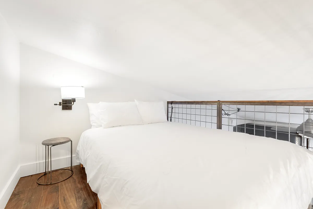 The image features a serene lofted bed with soft linens, a nightstand, wall sconce, and a railing. The subtle lighting and tranquil mood evoke comfort and privacy, emphasizing the bed as a restful retreat within the compact studio.