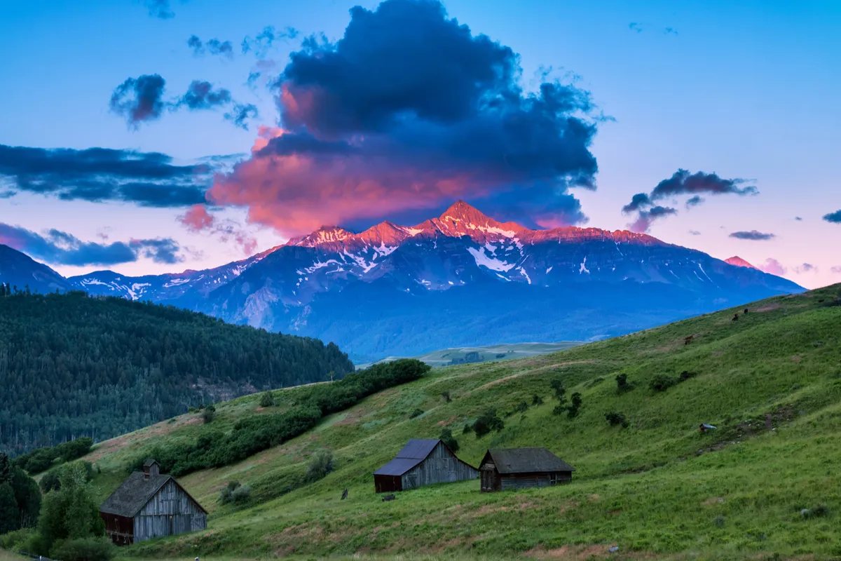 This exterior image features a tranquil mountain scene at sunset, with cozy cabins nestled in a meadow and the majestic peaks rising in the background. The soft clouds and fading light evoke a peaceful, restful atmosphere—an ideal setting for relaxation after a day of adventure.