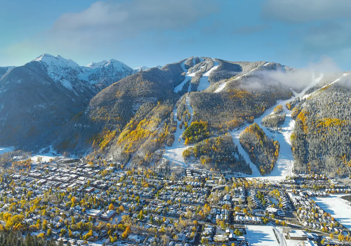 A sweeping view from above captures Telluride’s iconic snow-capped mountains, colorful autumn trees, ski slopes, and the lively town below. The image hints at the vibrant community atmosphere and seasonal celebrations that characterize the area.