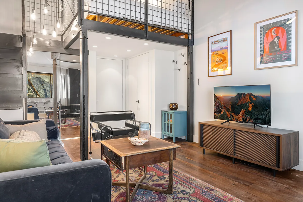 A cozy, contemporary living area featuring a plush sofa with colorful cushions, a wooden coffee table, a flat-screen TV displaying a mountain landscape, and artwork on the walls. Natural light fills the room, creating a welcoming atmosphere for relaxation and socializing.