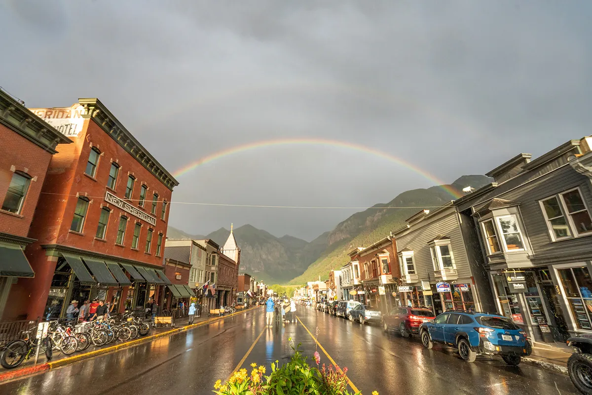 A vibrant scene on Telluride’s historic Main Street, featuring charming brick buildings, a rainbow arching overhead, and wet pavement suggesting a recent rain shower. Locals and visitors stroll along the sidewalks, some pausing to shop, dine at outdoor cafes, or take photos. The majestic mountains rise in the background, completing the quintessential small-town alpine vibe.