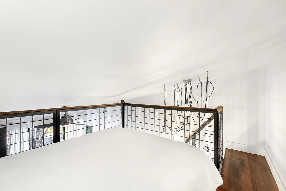 This image captures a lofted bed framed by a metal railing and wooden surfaces, with hanging wires adding to the loft’s character. The calm atmosphere suggests a peaceful spot for reading, relaxing, or sleeping, and the elevated position reinforces the loft’s secluded feel.