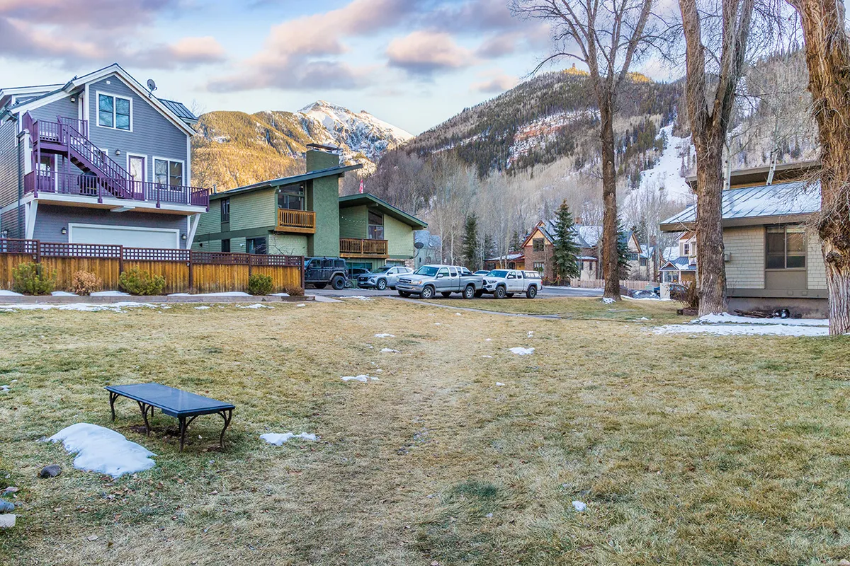 An outdoor space adjacent to a green building, framed by views of the mountains. The scene showcases a grassy community yard, where people can relax, gather, or park vehicles. The space feels open, sunny, and welcoming, ideal for social or solo lounging.