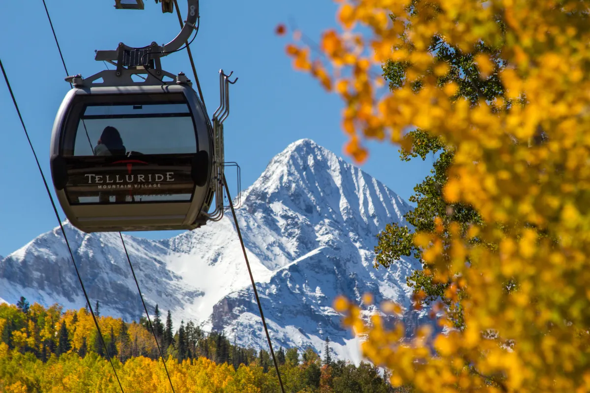 An exterior scene frames Telluride’s gondola crossing above autumn foliage and snow-capped mountains, representing easy access to outdoor adventures and town-wide festivities.