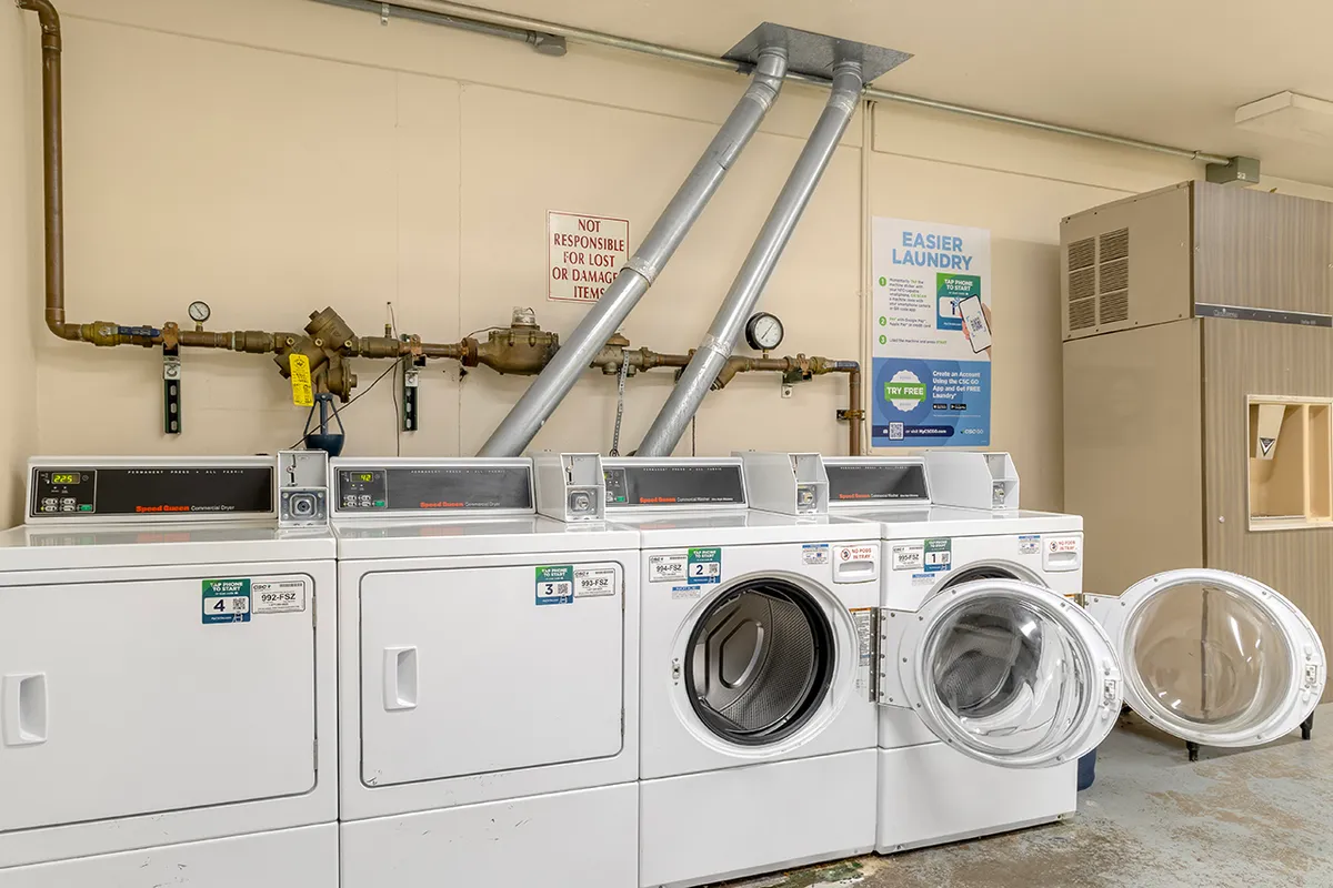 A functional, well-lit laundry room featuring multiple washing machines and dryers, with informational signage for user convenience. The arrangement suggests efficient, communal use and a sense of practicality.