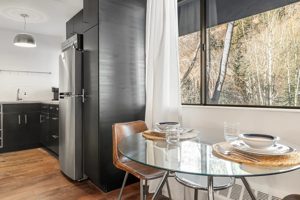 A cozy dining nook with a round glass dining table set beside a large window, modern kitchen appliances in the background, and natural sunlight streaming in. The space is perfect for enjoying meals or morning coffee while taking in the view.