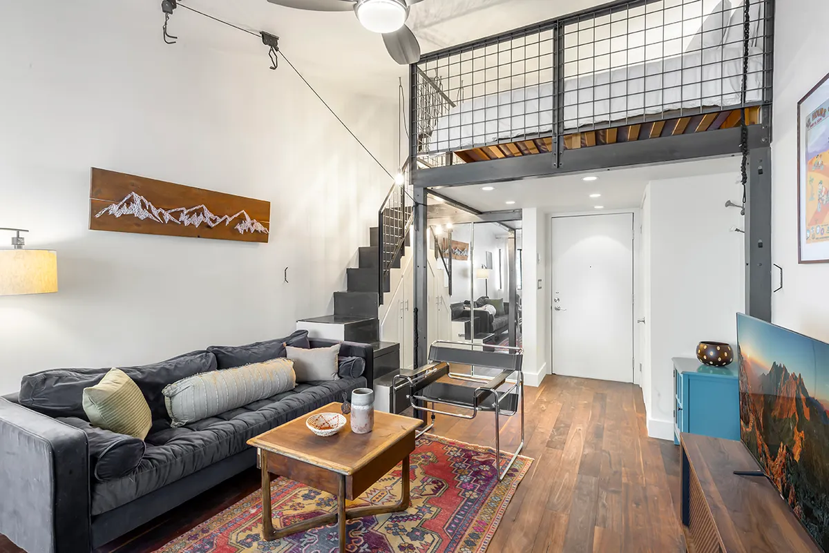 The image showcases a bright, stylish studio living area with a plush sofa, modern staircase leading to a cozy loft, eye-catching wall art, a sleek coffee table, and a mounted television. The space feels inviting and thoughtfully designed, perfect for relaxing or entertaining.
