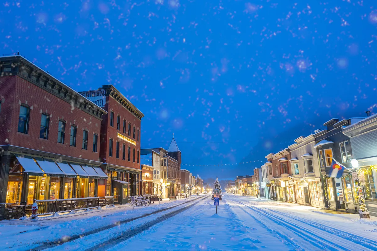 A festive evening view of Telluride’s Main Street adorned for the holidays. The snow-covered street is illuminated by shop window lights and a glowing Christmas tree, creating a magical atmosphere. Visitors stroll among the decorations, enjoying the seasonal charm and sense of community.