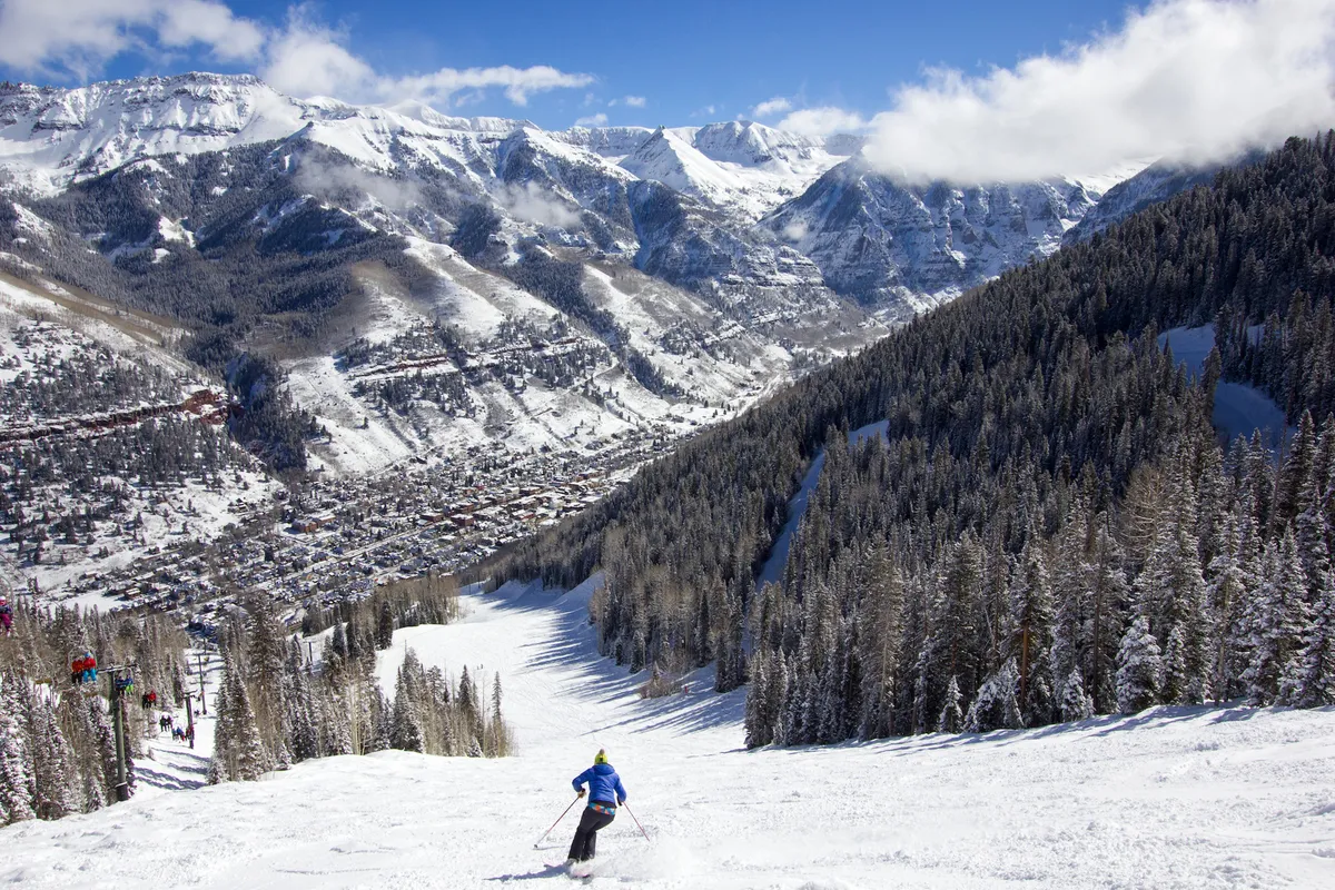 A skier carves down a snowy slope surrounded by majestic, snow-covered mountains and a dense pine tree line, with the charming town nestled in the valley below. The sky is clear, and the scene exudes energy and excitement, with the rugged natural landscape providing a stunning backdrop.