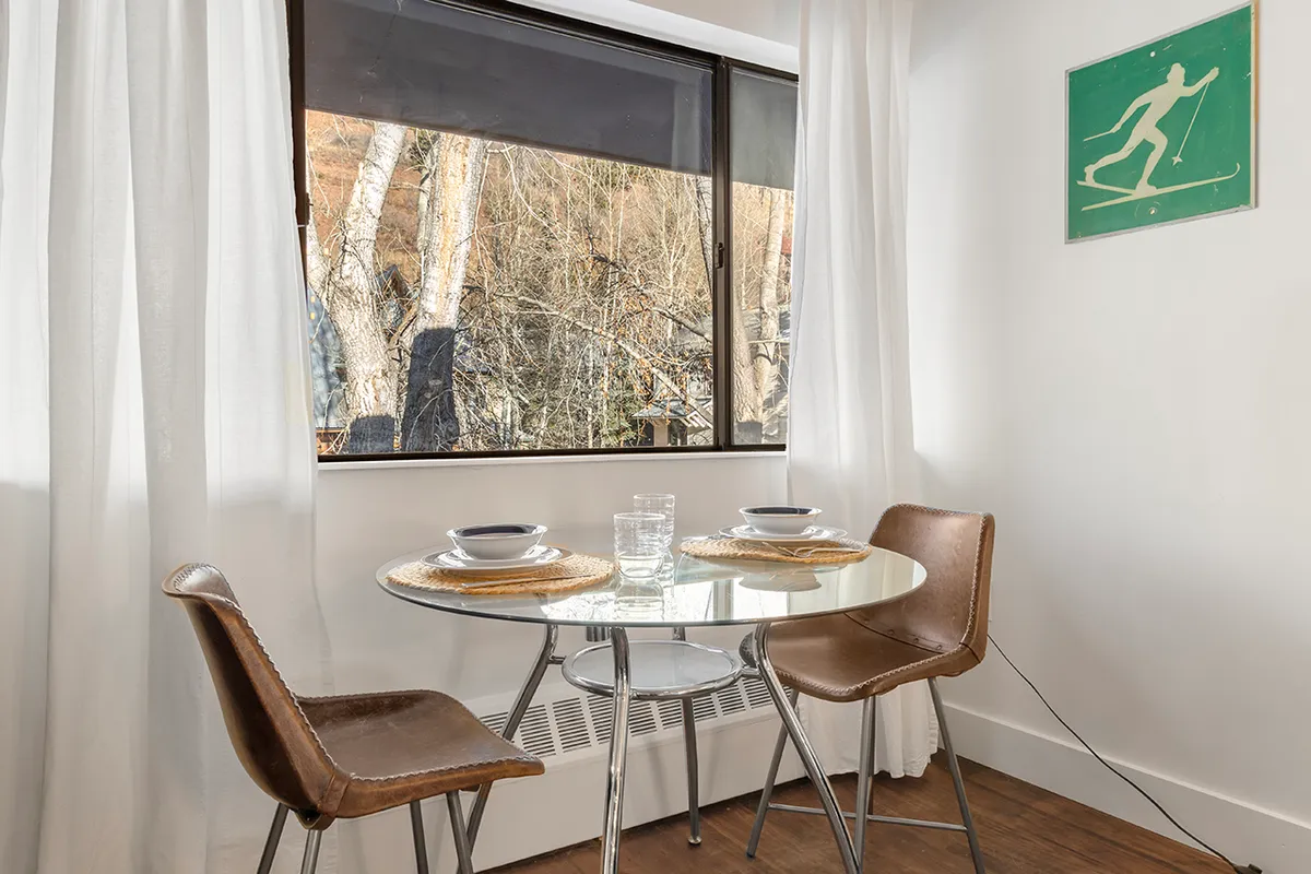 A window-side dining nook with a round glass dining table and two brown chairs, set against a large window offering a view. The space is accented by ski-themed artwork and bright, cheerful natural light, creating an ideal spot for intimate dining or morning coffee.