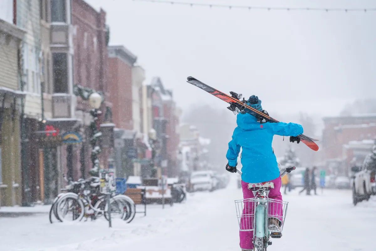 A snow-dusted street lined with colorful storefronts, where a person rides a bike equipped with skis. The scene exudes local charm and energy, with hints of winter activities and easy access to shops and cafés. The overall atmosphere is spirited and playful, emphasizing Telluride’s blend of outdoor adventure and small-town convenience.