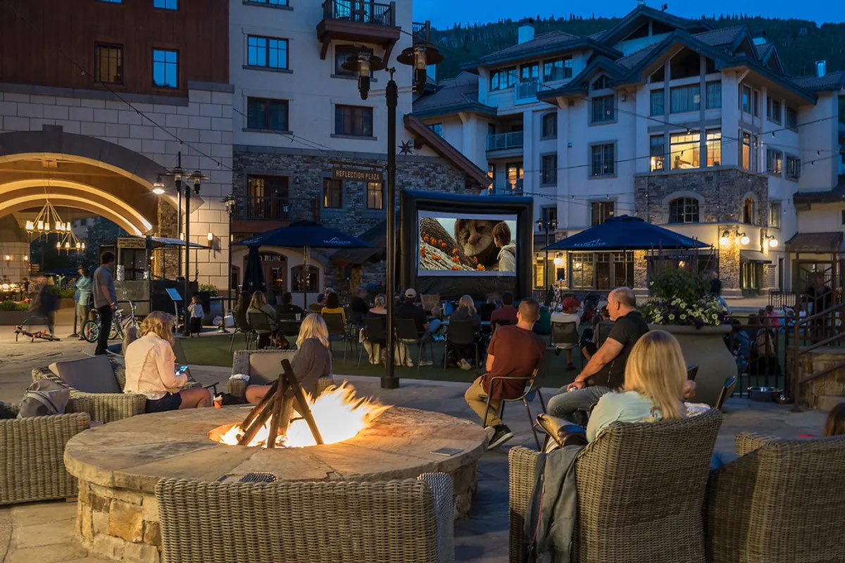 An inviting communal outdoor space with a fire pit, comfortable seating, and an outdoor movie screen, surrounded by mountain-inspired architecture. The scene is set for relaxing with friends or enjoying a quiet evening under the stars, reinforcing the communal yet cozy retreat theme.
