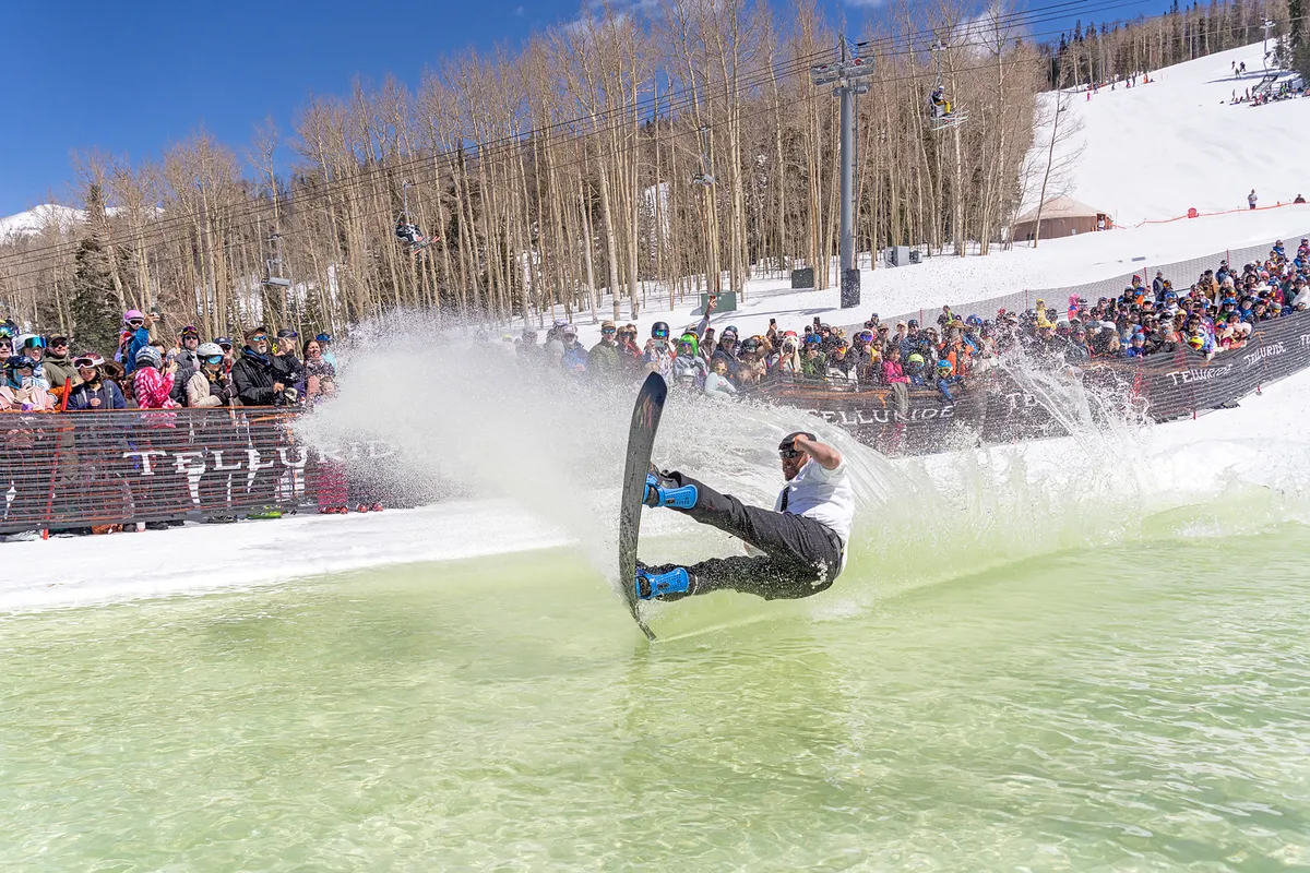 A dynamic winter event with a snowboarder splashing through a green water pond as a crowd of spectators, dressed in colorful ski attire, looks on. The scene is energetic, communal, and distinctly festive.