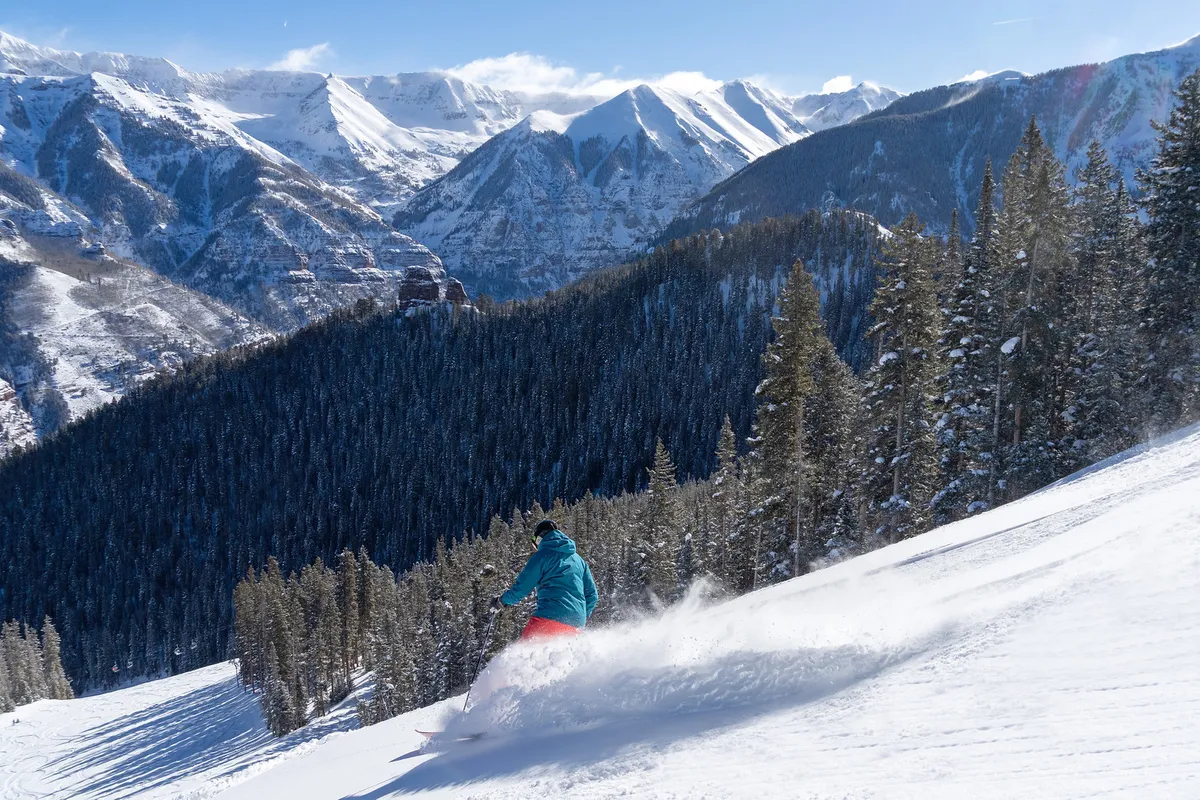 A snowboarder makes an exhilarating descent down a pristine, snow-covered mountain trail, flanked by snowy pine trees and expansive mountain vistas. The action-packed moment conveys the thrill and joy of winter sports.